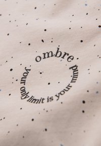 Světle béžová tkanina s černými a modrými skvrnami. Obsahuje kulaté textové pole: "ombre mind, your only limit is your." Text má tučný, moderní font.