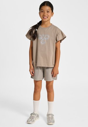 Hellbraunes Kurzarmhemd mit Rüschenärmeln und blauem grafischem Design, kombiniert mit hellgrauen Shorts und silbernen Turnschuhen.