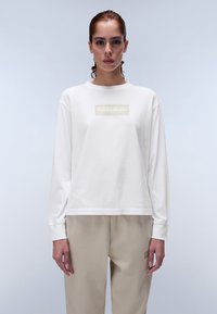 Valkoinen pitkähihainen t-paita, jossa on suorakulmainen beige logo rintakehässä, rento malli ja pehmeä puuvillamateriaali. Vaalea tausta.