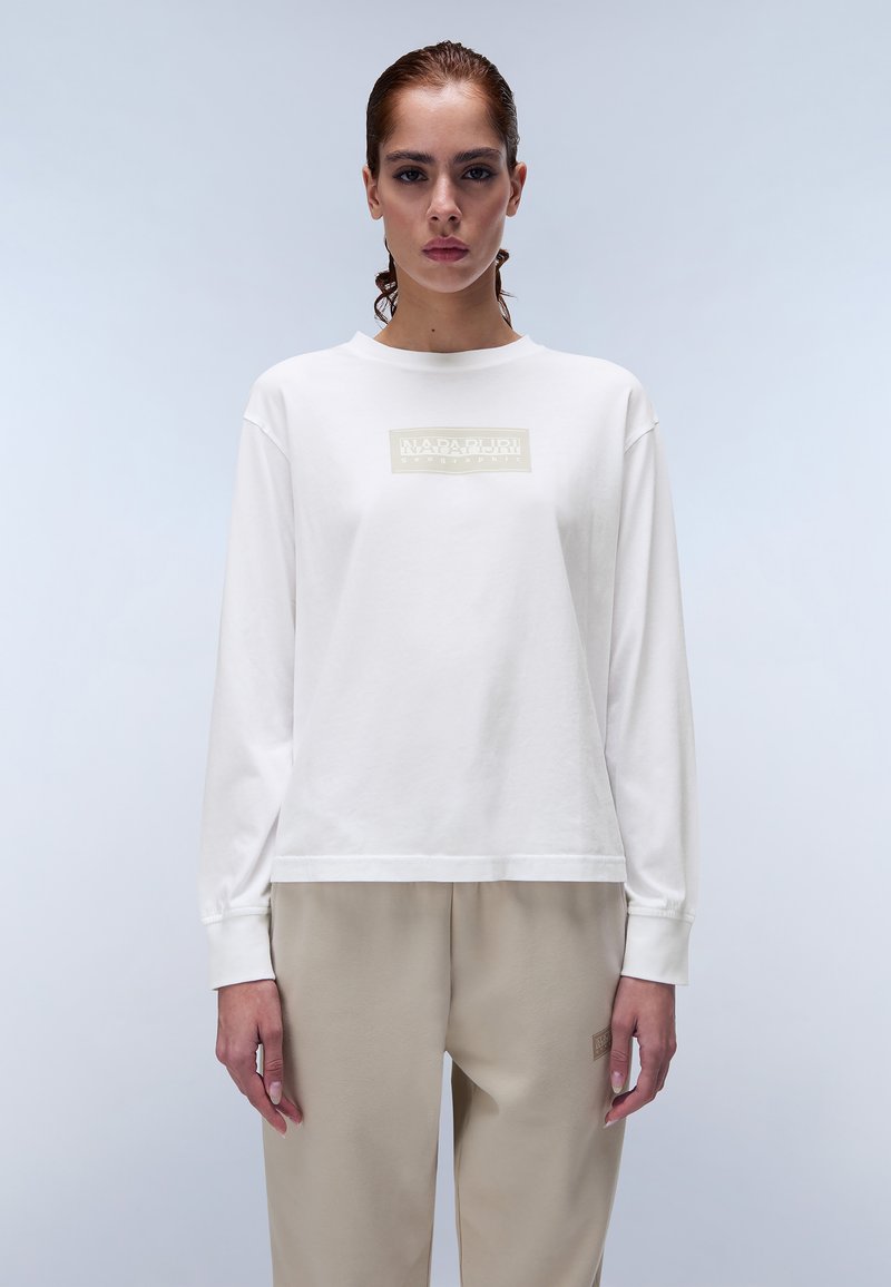 Valkoinen pitkähihainen t-paita, jossa on suorakulmainen beige logo rintakehässä, rento malli ja pehmeä puuvillamateriaali. Vaalea tausta.