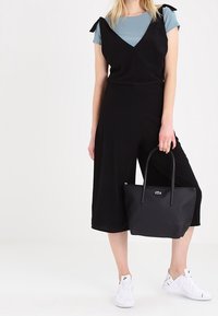Combinaison sans manches noire avec des accents de nœuds, associée à un t-shirt bleu clair, un sac à main noir et des baskets blanches. Tissu lisse et jambes raccourcies.