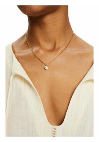 Collier en chaîne dorée ornée d'un pendentif rond avec une perle brillante. La personne porte un vêtement texturé de couleur claire avec un décolleté en V.
