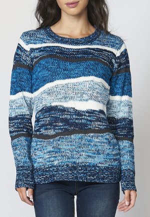 Femme portant un pull en maille à manches longues avec des rayures horizontales bleues, blanches et noires, associé à un jean bleu foncé.