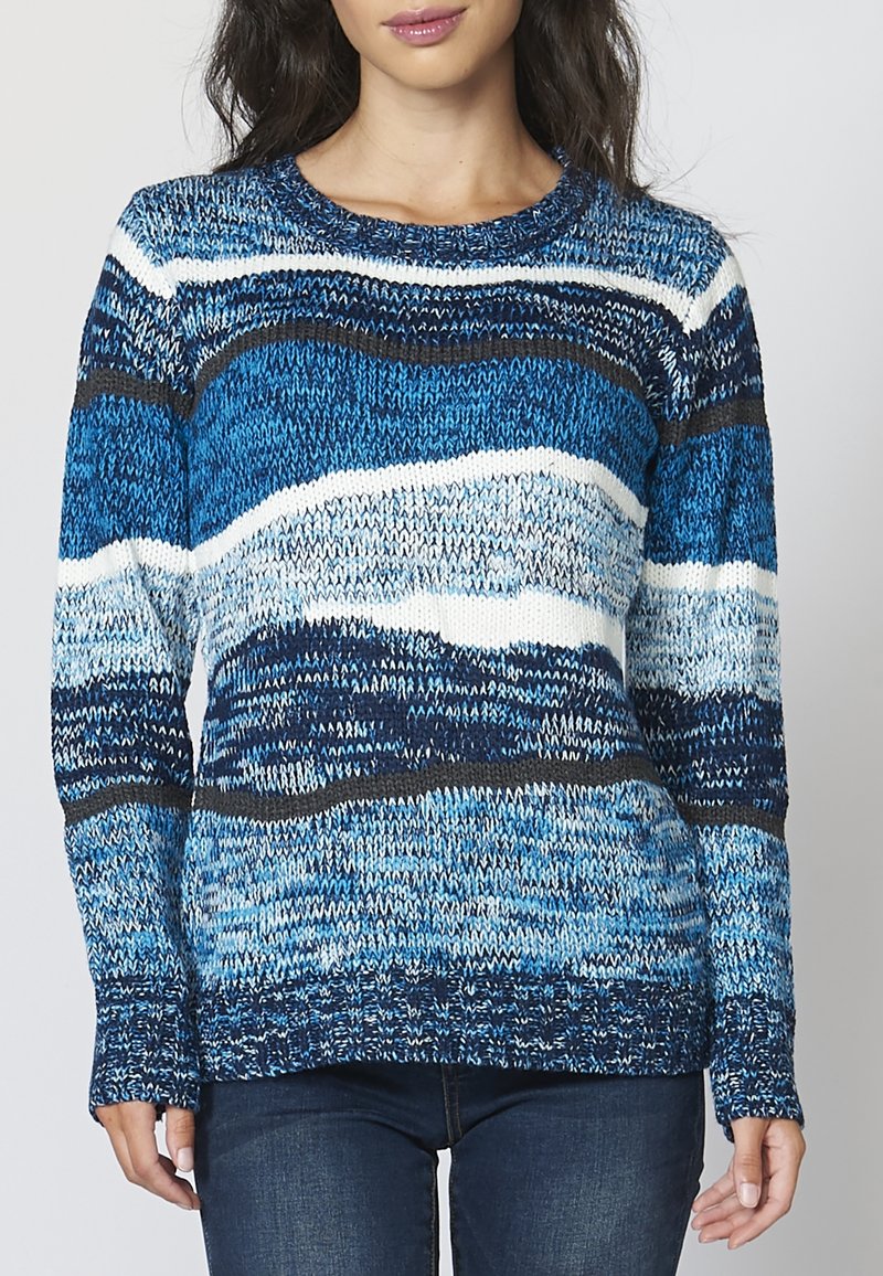 Femme portant un pull en maille à manches longues avec des rayures horizontales bleues, blanches et noires, associé à un jean bleu foncé.