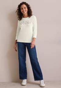 Light cream sweatshirt with "CREATE INSPIR" text, paired with dark blue wide-leg jeans and white sneakers. Smooth texture, casual fit.