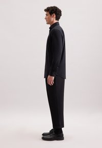 Camicia nera slim-fit con maniche lunghe, dotata di colletto e polsini con bottone, abbinata a pantaloni neri affusolati e scarpe nere.