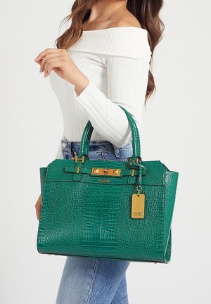 Woman holding green crocodile-texture leather handbag with gold hardware and branded tag, wearing white off-shoulder top and blue jeans.