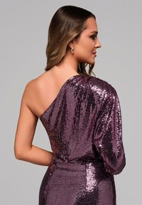 Lila paljettklänning med enaxlad design, figurnära siluett och texturerad yta. Lång ärm hänger elegant över armen.