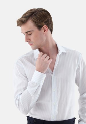 Junger Mann mit hellbraunem Haar, der ein weißes Hemd trägt, nach unten schaut und mit der rechten Hand seinen Kragen berührt.