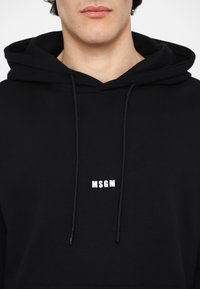 Homme portant un sweat à capuche noir avec le logo MSGM centré au-dessus de la poitrine, visible du cou au haut du torse sur fond blanc.