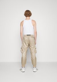 Beige cargo kalhoty s elastickými manžetami a bočními kapsami, vyrobené z hladké látky, v kombinaci s bílým tílkem, zobrazené zezadu.