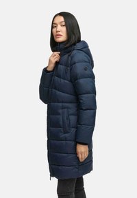 Mujer vistiendo un abrigo de invierno acolchado con capucha en azul marino, sujetando el cuello, de pie contra un fondo claro y liso.