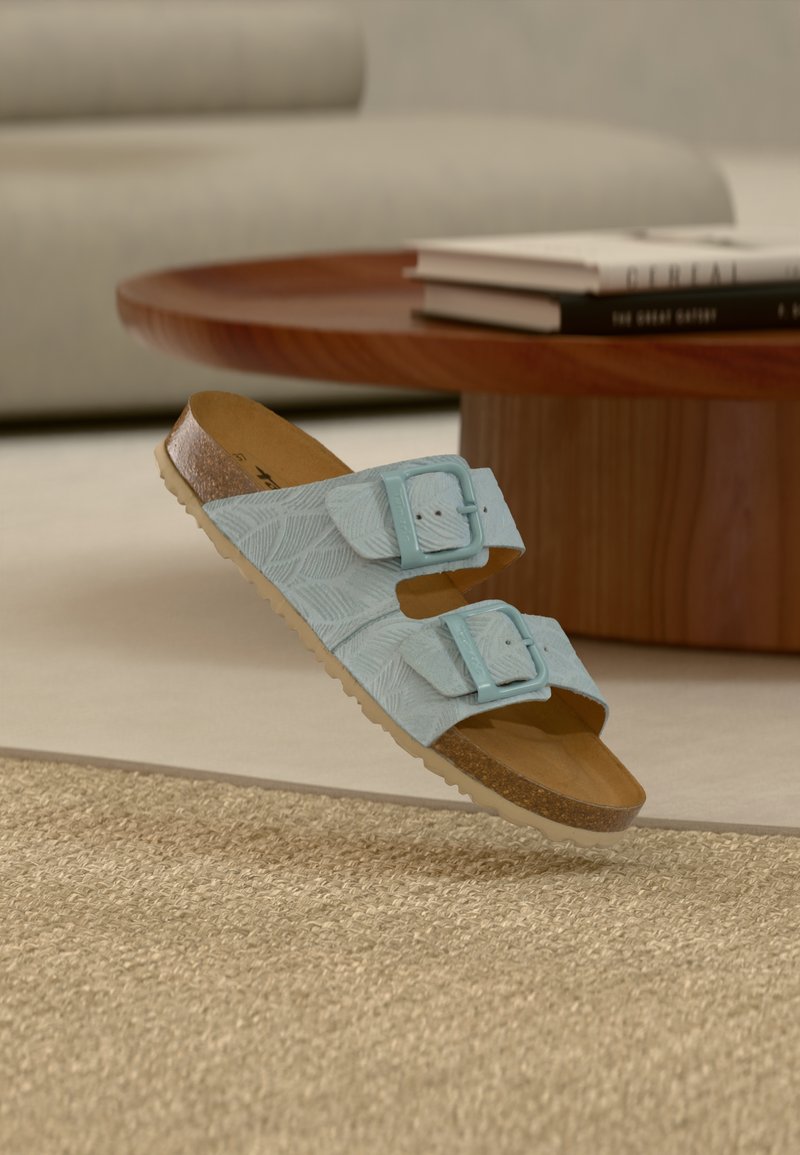 Sandales en cuir bleu clair avec deux brides à boucle, un motif texturé de feuilles, une semelle intérieure en liège et une semelle extérieure en caoutchouc blanc, flottant au-dessus d'un tapis.