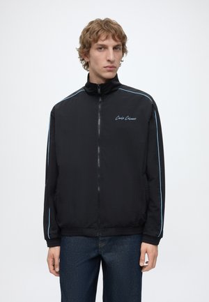 Jeune homme debout de face, portant une veste noire zippée avec des liserés bleus et le texte "Carlo Colucci" sur la poitrine, associé à un jean foncé.
