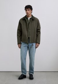 Man stands wearing dark olive jacket, green shirt, white undershirt, light blue jeans, and black shoes against a plain gray background.