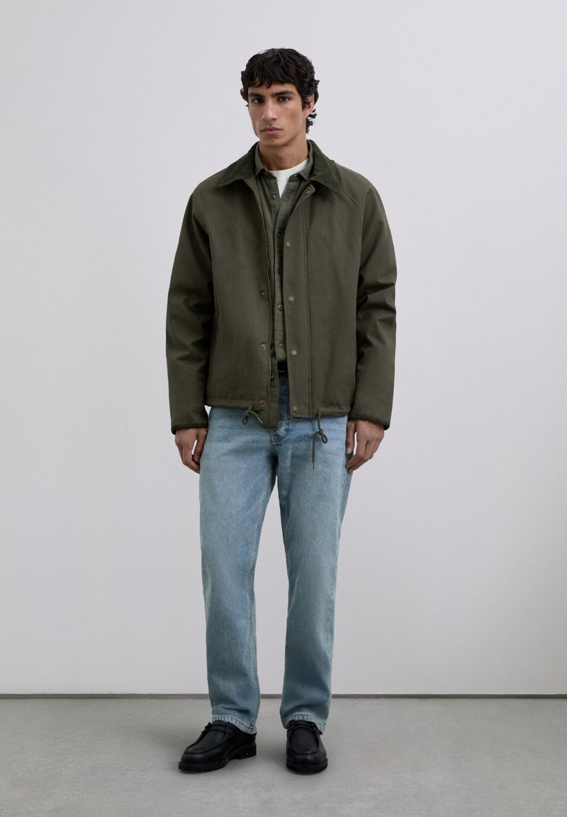 Man stands wearing dark olive jacket, green shirt, white undershirt, light blue jeans, and black shoes against a plain gray background.