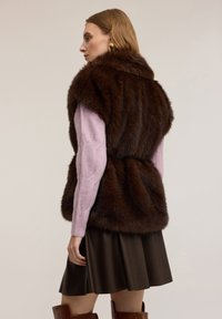 Kobieta ubrana w brązową futrzaną kamizelkę na lawendowym swetrze, ciemnobrązową skórzaną spódnicę i brązowe kozaki do kolan, stojąca bokiem na jednolitym tle.