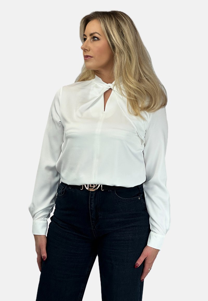 Biała bluzka z długimi rękawami z dekoltem typu keyhole, zestawiona z ciemnoniebieskimi spodniami z wysokim stanem i błyszczącym paskiem. Gładka faktura materiału.