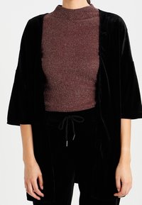 Femme portant un haut à col roulé maroon pailleté sous une veste ouverte en velours noir ample à manches trois-quarts.