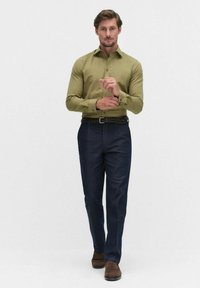 Olijfgroene long sleeve shirt, donkerblauwe broek, gevlochten zwarte riem, lichtbruine schoenen. Model staat rechtop met de handen samengevouwen.