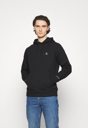 Junger Mann trägt schwarzen Calvin Klein Hoodie mit Logo auf Brust und Ärmel, kombiniert mit blauen Jeans, steht vor weißem Hintergrund.