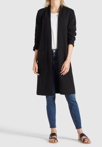 Femme portant un long cardigan noir ouvert, un t-shirt blanc, un jean skinny bleu et des sandales noires, debout devant un fond uni.