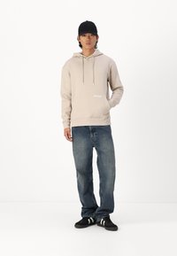 Felpa beige con cappuccio, cordini e tasca frontale, abbinata a jeans in denim blu larghi e scarpe nere con strisce bianche.