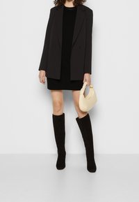 Femme portant un blazer noir, une mini robe noire, des bottes noires jusqu'aux genoux, et tenant un petit sac à main beige sur un fond uni.