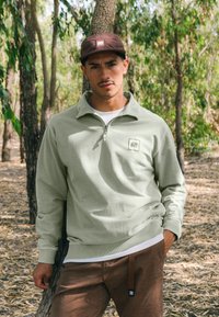 Sudadera con cremallera de color verde claro, con puños y dobladillo acanalados, que cuenta con un pequeño parche de logo, combinada con pantalones marrones y llevada en un entorno forestal.