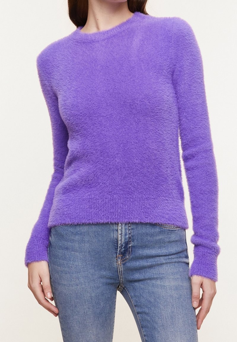 Femme portant un pull violet ajusté à manches longues en tissu duveteux, associé à un jean en denim bleu, sur un fond uni.