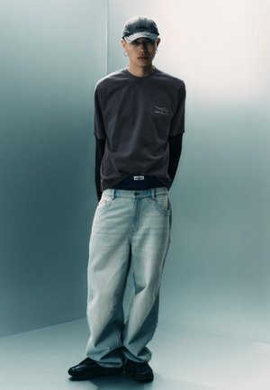 Mladić u sivoj kapici, prevelikoj tamnoj majici preko dugih rukava, svijetlim širokim trapericama i crnim cipelama stoji naslonjen na jednobojni zid.