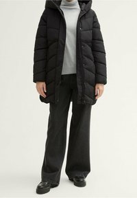 Abrigo acolchado negro con capucha, con un diseño de acolchado, bolsillos laterales y cierre de cremallera, usado sobre un jersey gris de cuello alto y jeans de pierna ancha.