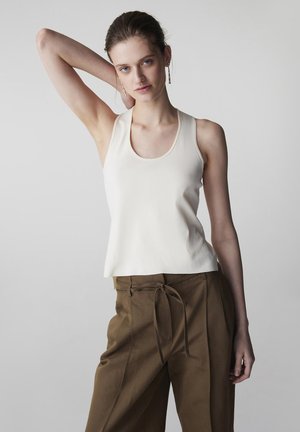 Joven posando con un brazo levantado detrás de la cabeza, vistiendo una blusa blanca sin mangas y pantalones marrones con cordón, sobre un fondo liso.