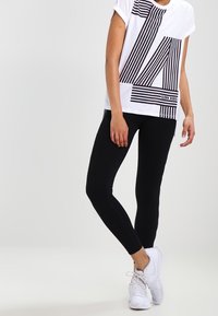 Hvid t-shirt med korte ærmer og sorte striber, der danner et '14' design og logo, parret med sorte leggings og hvide sneakers.