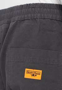 Pantaloni de lucru gri închis dintr-un material durabil, cu talie elastică și o etichetă dreptunghiulară portocalie cu inscripția "Service Works".