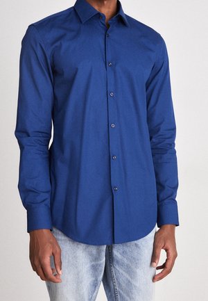 Mann trägt ein enganliegendes, langärmeliges, blaues Hemd mit Knöpfen, kombiniert mit hellblauen Jeans, vor einem schlichten Hintergrund.