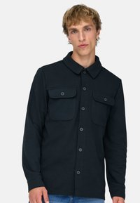 Junger Mann mit blondem Haar, der ein schwarzes Hemd mit Klappentaschen und blaue Jeans trägt, vor einem einfarbigen Hintergrund stehend.