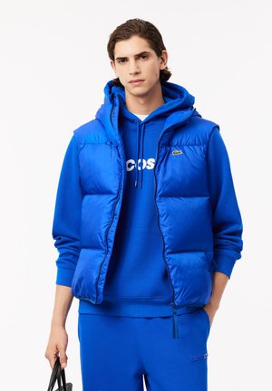 Blau gepolsterte Weste mit Kapuze, die eine glatte Textur, einen Reißverschluss und ein leichtes Design aufweist, getragen über einem passenden blauen Sweatshirt.