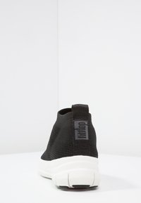 Zwarte slip-on sneaker met een gestructureerde mesh bovenkant, witte zool en een treklip met het "fitflop" logo aan de achterkant.