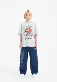 Light gray oversized t-shirt featuring a colorful skull and heart graphic. Paired with loose blue denim jeans adorned with beaded "Ed Hardy" text.