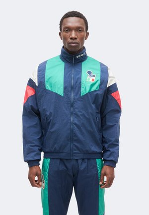Muž oblečený v tmavomodro-zeleno-červenom súprave Adidas so zipsovou bundou, ktorá má nášivku s hviezdami a talianskou vlajkou.