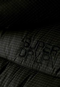 Svart tyg med ett rutmönster som har broderad text "SUPER DRY" i mörk tråd, vilket visar en slät textur och modern design.