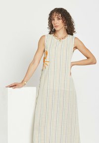 Mujer de cabello rizado que lleva un vestido largo sin mangas a rayas con bordados naranjas, de pie con una mano en la cadera y la otra apoyada en un pedestal blanco.