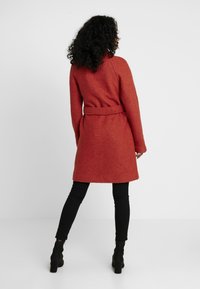 Röd ullkappa med skärp, knälång, figursydd silhuett, texturerat tyg, långa ärmar, bärs med svarta figursydda byxor och ankelboots.