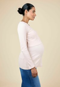 Hea roosa soonikkoiga pikkade varrukatega pluus, mis on keha allpool keha ümber kinni, ümarate servadega, koos siniste teksadega. Küljelt vaade rasedale naisele.