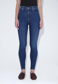 Jeans de mezclilla azul de cintura alta con un corte ajustado, que incluyen bolsillos frontales y un cierre de cremallera clásico. Textura suave con mínimas costuras.