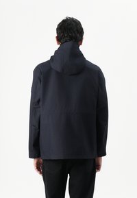 Marineblå hættejakke med en afslappet pasform, der har en glat tekstur og skuldersøm-detaljer. Sammen med mørke denim bukser.