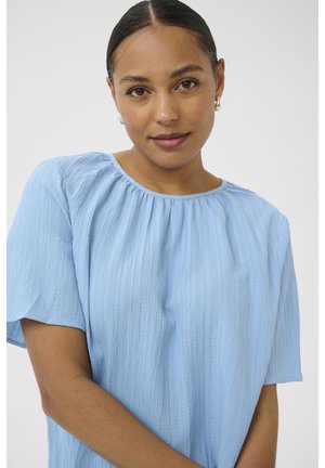 Nainen, jolla on tummat hiukset yhteen sidottuina, pukeutuneena vaaleansiniseen teksturoituun lyhythihaiseen puseroon ja hopeisiin renkaskorvakoruihin, katsomassa eteenpäin neutraali ilme kasvoillaan.