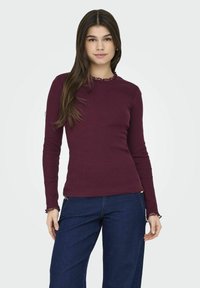 Top bordeaux a maniche lunghe in tessuto a coste con scollatura ondulata e dettagli ai polsini, abbinato a pantaloni in denim blu a gamba larga.