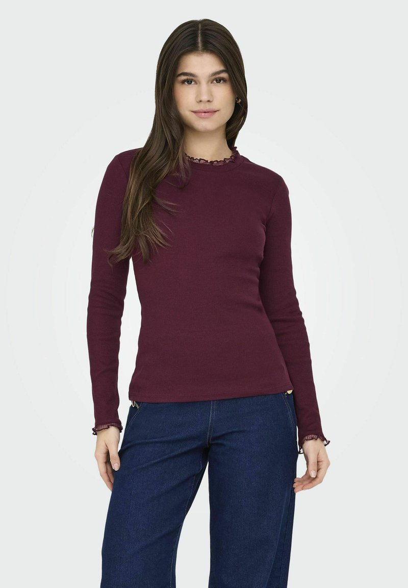 Top bordeaux a maniche lunghe in tessuto a coste con scollatura ondulata e dettagli ai polsini, abbinato a pantaloni in denim blu a gamba larga.
